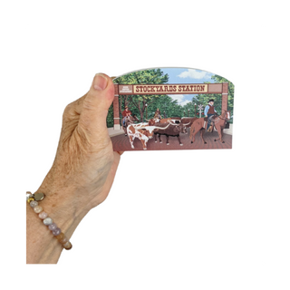 Hand holding a 3/4" thick wooden cutout  with a 'Stockyards Station' illustration featuring cowboys and cattle.