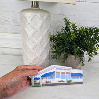 Hand holding a miniature model of a building in front of a decorative lamp and plant.