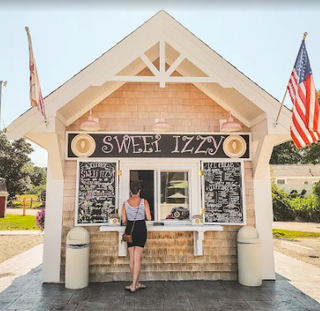 Small wooden building with 'Sweet Izzy' sign, person standing outside, American flags on either side.