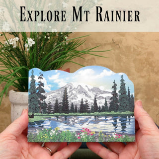 3/4" wooden cut out replica of a scenic illustration of Mount Rainier held by hands against a natural background.