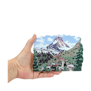 Hand holding a 3/4" thick wooden replica with an Illustration of a mountain and village illustration