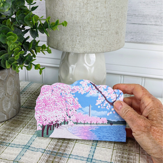 Hand holding a 3/4" thick wooden illustration of cherry blossom trees and the Washington Monument with a lake, placed on a table with a lamp and plant in the background.