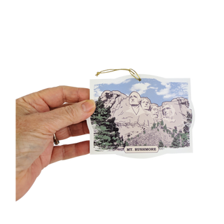 Hand holding a 1/4" thick pvc ornament  with Mount Rushmore illustration on a white background