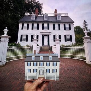 A miniature wooden model of a white house with black shutters is held up by a hand against a real background of a large white historic house on a brick driveway.