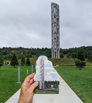 A hand holds a wooden replica showing a tall concrete monument over a grassy landscape, with the real monument visible in the background outdoors under a cloudy sky.