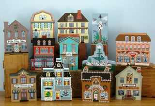 A collection of handcrafted, colorful miniature wooden buildings and storefronts arranged on a wooden surface against a light blue background.