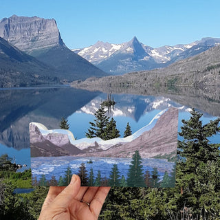 A hand holds a small printed card or photo showing a mountain lake landscape with snowy peaks and pine trees, placed in front of a much larger matching landscape photo of the same location.